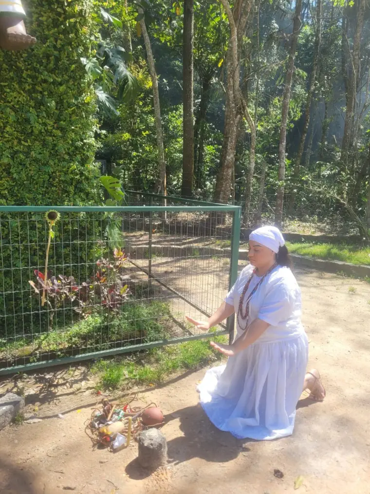 Yalorixá Cigana ajoelhada na mata, com as mãos estendidas sobre uma oferenda no chão