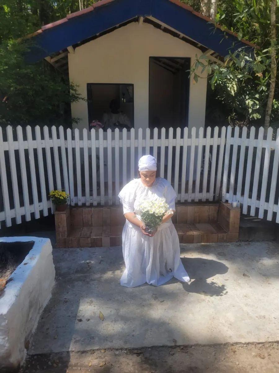 Yalorixá Cigana de joelhos, toda de branco, segurando um vaso de margaridas brancas diante de uma casinha de reza