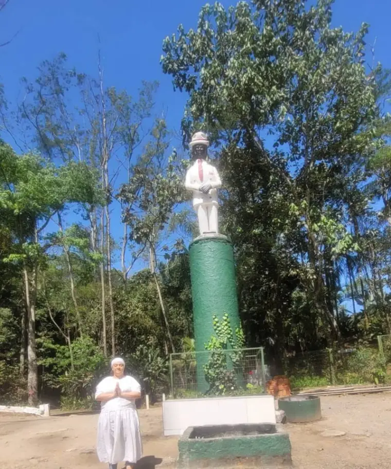 Yalorixá Cigana de mãos postas, toda de branco, diante de uma imagem no alto de uma coluna verde